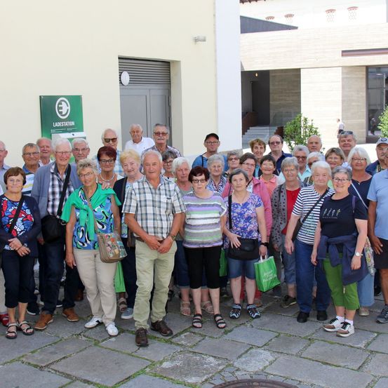 Eine Gruppe von Menschen steht zusammen vor einem Gebäude. Sie sind alle lässig gekleidet, einige tragen Brillen und einige halten Taschen. Ein grünes Schild mit dem Wort Ladestation ist an der Wand.