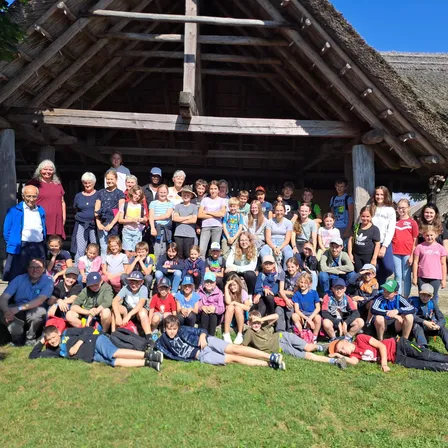 Eine große Gruppe von Menschen, darunter Erwachsene und Kinder, versammelt sich vor einem strohgedeckten Gebäude. Sie lächeln und posieren für ein Foto. Einige sitzen auf dem Gras, während andere stehen.