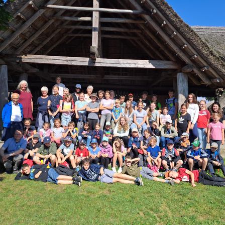 Eine große Gruppe von Menschen, darunter Erwachsene und Kinder, versammelt sich vor einem strohgedeckten Gebäude. Sie lächeln und posieren für ein Foto. Einige sitzen auf dem Gras, während andere stehen.