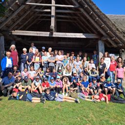 Eine große Gruppe von Menschen, darunter Erwachsene und Kinder, versammelt sich vor einem strohgedeckten Gebäude. Sie lächeln und posieren für ein Foto. Einige sitzen auf dem Gras, während andere stehen.