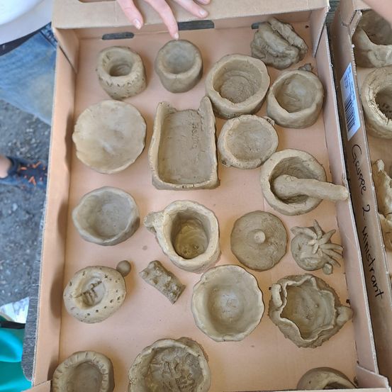 Eine Schachtel enthält verschiedene Gegenstände aus Ton, darunter Schalen, einen Mörser und kleine Skulpturen. Der Ton ist unbemalt, und die Gegenstände variieren in Größe und Form.