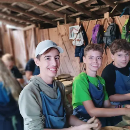Drei Jungen in Schürzen lächeln für ein Foto in einer Werkstatt. Holzwände und hängende Rucksäcke im Hintergrund.