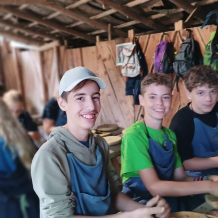 Drei Jungen in Schürzen lächeln für ein Foto in einer Werkstatt. Holzwände und hängende Rucksäcke im Hintergrund.