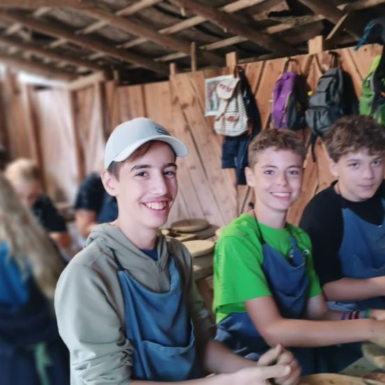 Drei Jungen in Schürzen lächeln für ein Foto in einer Werkstatt. Holzwände und hängende Rucksäcke im Hintergrund.