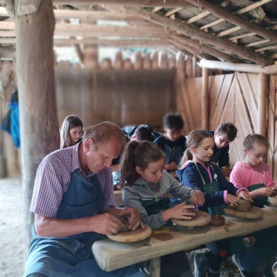 Eine Gruppe von Kindern sitzt um einen Holztisch in einer Scheune und trägt Schürzen. Ein erwachsener Mann lehrt sie Töpferwaren herzustellen.