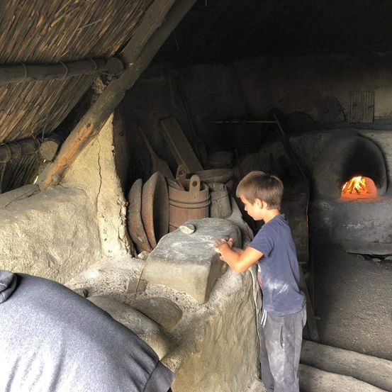 Ein junger Junge steht in einem Raum mit einer Steinmauer, hält einen Stein und es gibt einen Steinofen mit Feuer darin.