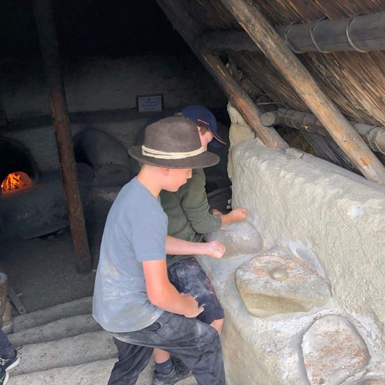 Zwei Jungen klettern eine Steinmauer hinauf, einer trägt einen Hut und der andere eine blaue Kappe. Hinter ihnen befindet sich eine Steinstruktur mit einer offenen Tür und einem Feuerloch.
