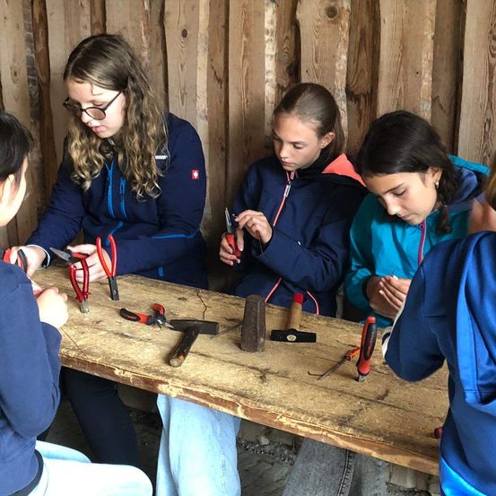 Eine Gruppe junger Mädchen sitzt um einen Holztisch herum mit verschiedenen Werkzeugen. Sie scheinen an einem Projekt zu arbeiten. Sie tragen alle Jacken und einige haben eine Brille. Das Mädchen auf der linken Seite hält eine Schere.