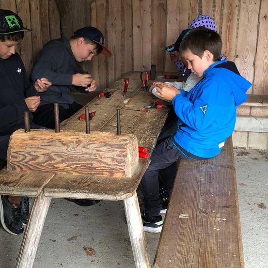 Eine Gruppe von Kindern sitzt um eine Holzbank herum und ist mit einer praktischen Aktivität beschäftigt. Ein Kind konzentriert sich auf ein Werkzeug, während andere zuschauen. Werkzeuge und Holzbretter liegen auf dem Tisch.