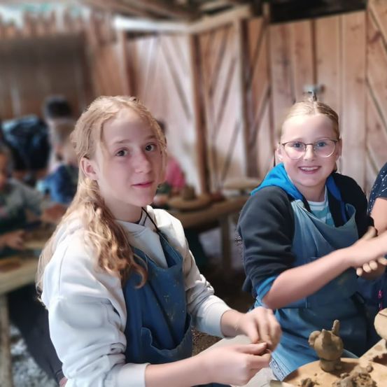 Zwei junge Mädchen arbeiten in einem Raum mit Holzwänden an Töpferwaren. Ein Mädchen trägt ein weißes Hemd und eine Brille. Hinter ihnen arbeitet eine Person an Ton. Der Raum ist schwach beleuchtet.