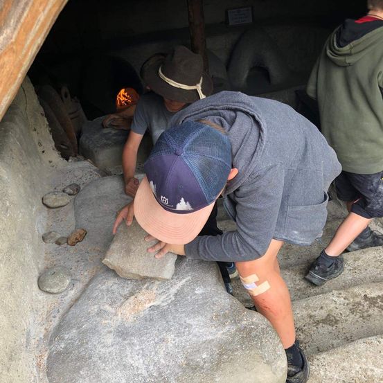 Zwei Personen arbeiten an einem Felsen in einer Steinstruktur, einer benutzt ein Werkzeug. Im Hintergrund brennt ein Feuer.