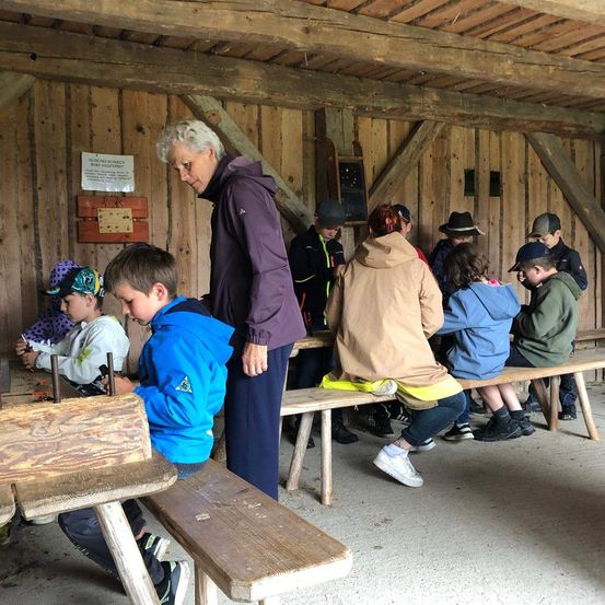 Eine Gruppe von Kindern sitzt auf Bänken in einem Holzraum, mit einer Frau vor ihnen. Sie machen einige Aktivitäten, und einige von ihnen tragen Mützen und Turnschuhe.
