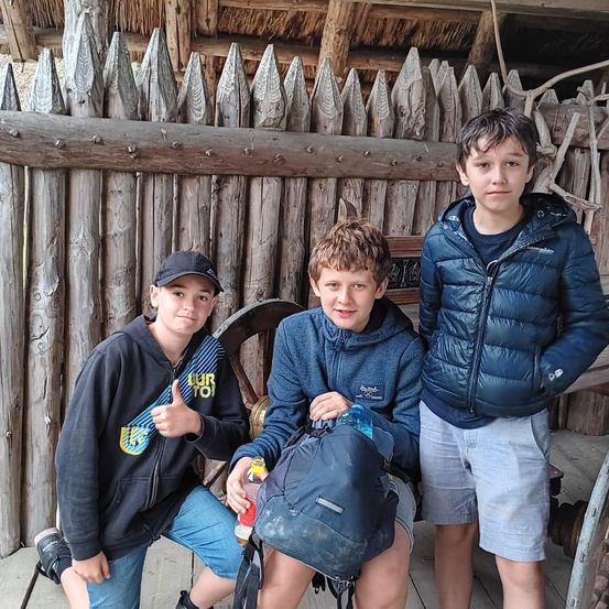 Drei Jungen posieren für ein Foto. Sie stehen vor einem Holzzaun mit einem Rad. Der Junge in der Mitte sitzt und lächelt. Der Junge rechts steht mit den Händen in den Taschen. Der Junge links gibt einen Daumen hoch.