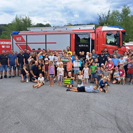 Eine große Gruppe von Erwachsenen und Kindern posiert vor einem Feuerwehrauto. Sie halten Geschenke und lächeln für ein Foto.