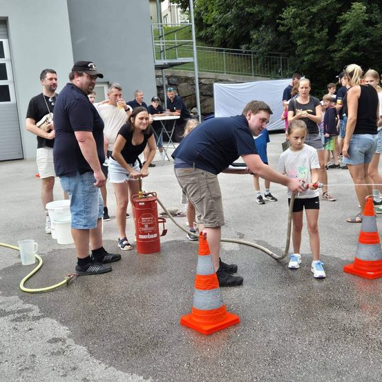 Eine Gruppe von Menschen versammelt sich um einen Feuerlöscher, ein Mann füllt ihn mit Wasser, während ein junges Mädchen neben ihm steht.