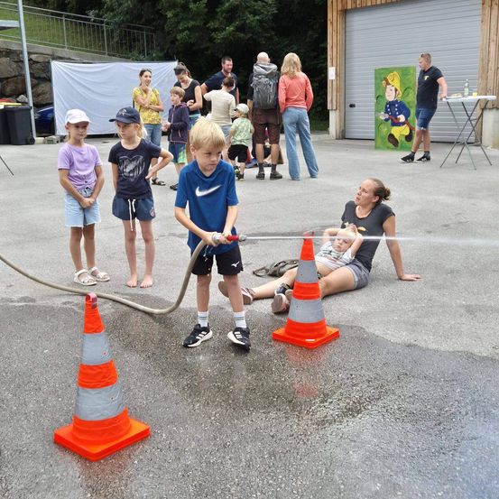 Kinder spielen mit einem Wasserschlauch und sprühen Wasser, während andere zusehen. Ein Erwachsener liegt mit einem Baby auf dem Boden. Verkehrskegel stehen auf der nassen Straße.