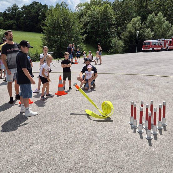 Eine Gruppe von Kindern und Erwachsenen nimmt an einer Feuerwehrübung auf einem gepflasterten Bereich mit Verkehrskegeln und einem Feuerwehrwagen im Hintergrund teil.