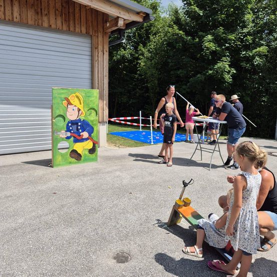 Eine Gruppe von Kindern und Erwachsenen ist draußen. Ein Mädchen sitzt auf einer Wippe. In der Nähe steht ein Gemälde eines Feuerwehrmanns auf dem Boden. Ein Tisch mit Essen und einer Frau befindet sich im Hintergrund.