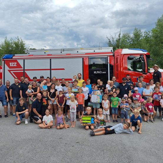 Eine große Gruppe von Menschen, einschließlich Erwachsener und Kindern, posiert für ein Foto vor einem roten Feuerwehrauto. Sie halten Geschenke und einige tragen Hüte.