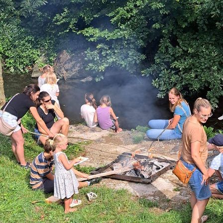Eine Gruppe von Menschen sitzt um ein Lagerfeuer herum und brät Essen. Einige sitzen auf dem Boden, andere stehen. Sie sind von Grün umgeben und von einem fließenden Fluss.