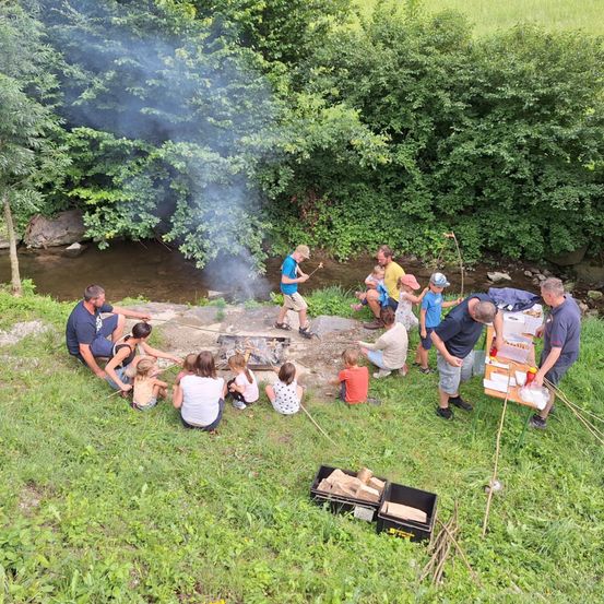 Eine Gruppe von Menschen ist um ein Lagerfeuer in einer Lichtung versammelt, mit einem Bach in der Nähe. Einige sitzen, andere stehen, und es gibt einen Tisch mit Essen.