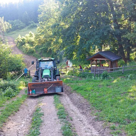 Ein Traktor steht auf einem Feldweg mit einem Holzzaun und einem Schuppen. In der Nähe sind Menschen, die möglicherweise ein Feuer pflegen. Das Gebiet ist von Bäumen und Grünflächen umgeben.