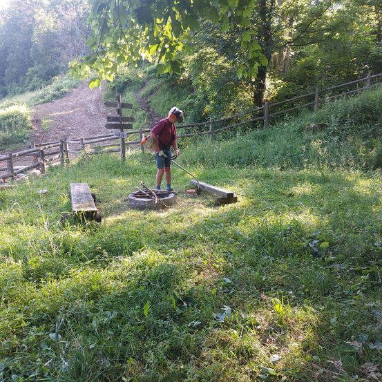 Ein Mann mit Kopfhörern und einem Rucksack verwendet einen Rasentrimmer, um das Gras um ein Feuerloch in einem Grasbereich zu schneiden. Ein Holzbank und ein Holzzaun sind in der Nähe.