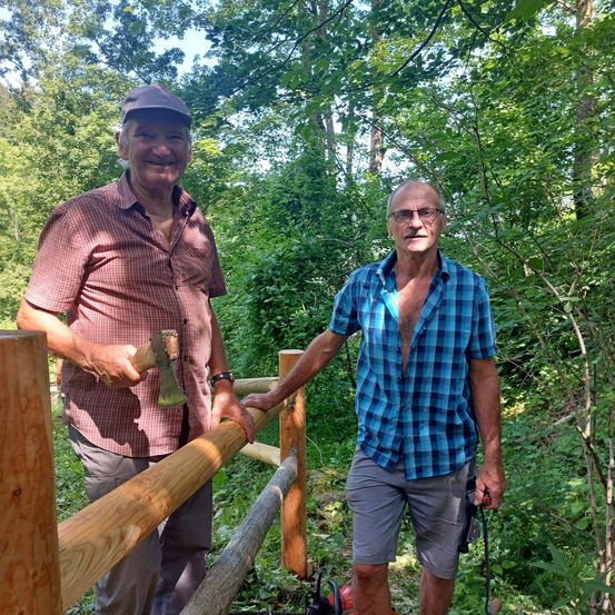 Zwei ältere Männer stehen auf einem Holzzaun in einem Wald. Einer hält eine Axt und der andere hat einen Hund an der Leine.