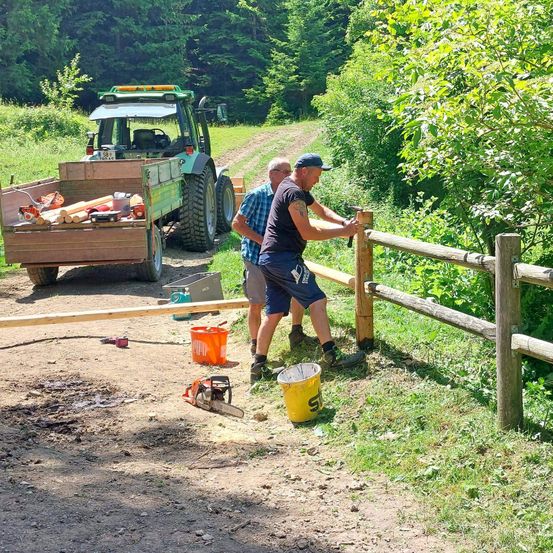 Zwei Männer reparieren einen Holzzaun auf einer unbefestigten Straße, hinter ihnen steht ein Traktor. Einer nagelt einen Pfosten, während der andere zusieht. Werkzeuge und Eimer sind herumliegen.