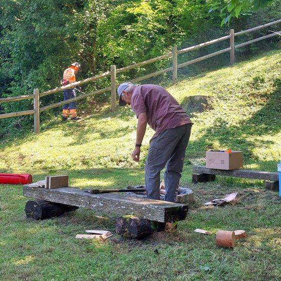 Ein Mann arbeitet im Freien und beugt sich über eine Holzplatte. In der Nähe bedient eine weitere Person in Sicherheitsausrüstung ein Werkzeug. Ein Holzzaun, Bäume und ein rotes Objekt sind im Hintergrund zu sehen.