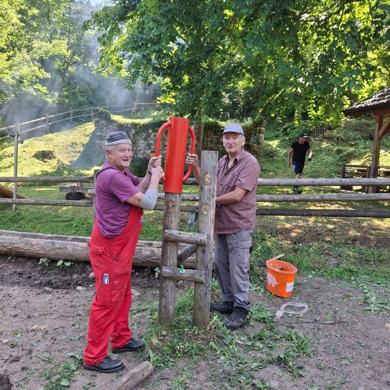 Zwei Männer im Freien, einer repariert ein rotes Objekt an einem Holzpfosten, der andere hält eine Holzkonstruktion. Ein orangefarbener Eimer und ein Mann, der im Hintergrund geht. Bäume und ein Holzzaun.