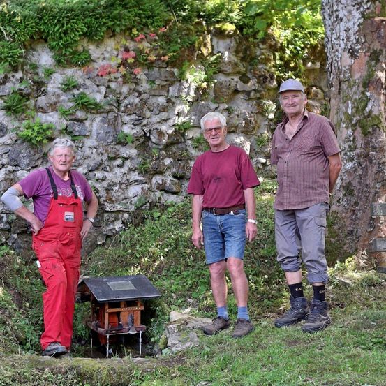Drei Männer stehen draußen in der Nähe einer Steinmauer. Der Mann auf der linken Seite trägt eine rote Latzhose. Der Mann in der Mitte trägt ein kastanienbraunes Hemd und blaue Shorts. Der Mann auf der rechten Seite trägt ein gestreiftes Hemd und graue Hosen.