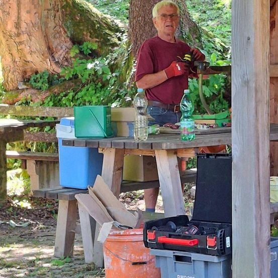 Ein älterer Mann mit Handschuhen arbeitet an einem Holztisch im Garten. Verschiedene Gegenstände wie Flaschen und ein Werkzeugkasten liegen auf dem Tisch. Ein Baumstamm und Pflanzen sind im Hintergrund.