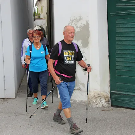 Eine Gruppe älterer Personen mit Trekkingstöcken geht durch einen schmalen Durchgang, zwei Männer und zwei Frauen. Sie gehen vor einem Gebäude mit einer grünen Tür.