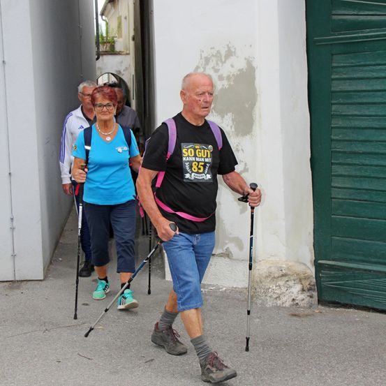 Eine Gruppe älterer Personen mit Trekkingstöcken geht durch einen schmalen Durchgang, zwei Männer und zwei Frauen. Sie gehen vor einem Gebäude mit einer grünen Tür.