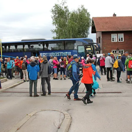 Eine Menschenmenge, einige mit Rucksäcken, geht um einen blauen Bus mit der Aufschrift 'EDTBRUSTNER' herum, in der Nähe eines Hauses mit braunem Dach.