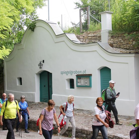 Eine Gruppe von Wanderern geht an einem weißen Gebäude mit einer grünen Tür, grünen Fensterläden und einer Website-Adresse über der Tür vorbei. Bäume und eine Steinmauer sind im Hintergrund.