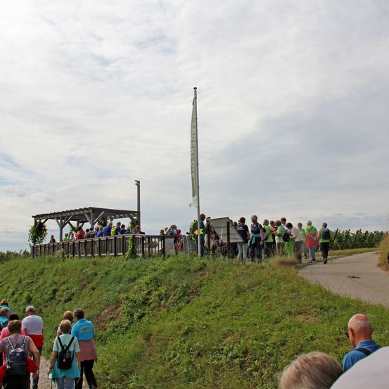 Eine Gruppe von Menschen geht auf einem Pfad in einem Grasbereich mit einem Holzzaun und einem Mast mit einer Fahne. Einige von ihnen tragen Rucksäcke. In der Ferne befindet sich ein Gebäude mit einem Dach. Der Himmel ist bewölkt.
