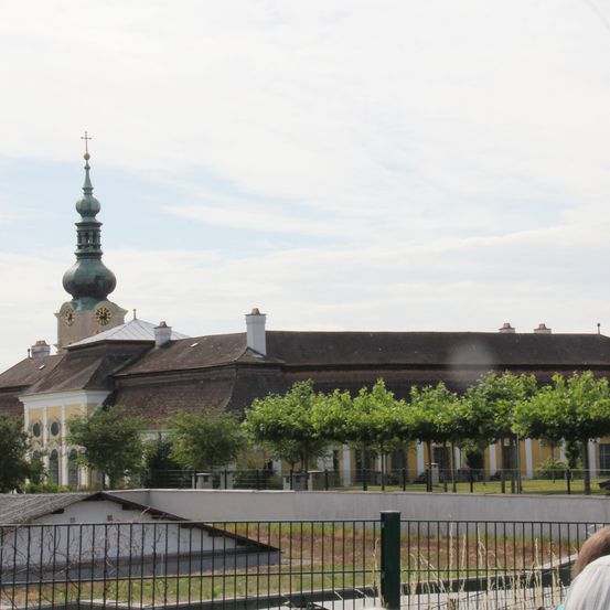 Ein großes Gebäude mit einem grünen Turm und einer Uhr, umgeben von Bäumen und einem Zaun. Das Gebäude hat ein braunes Dach und gelbe Wände.