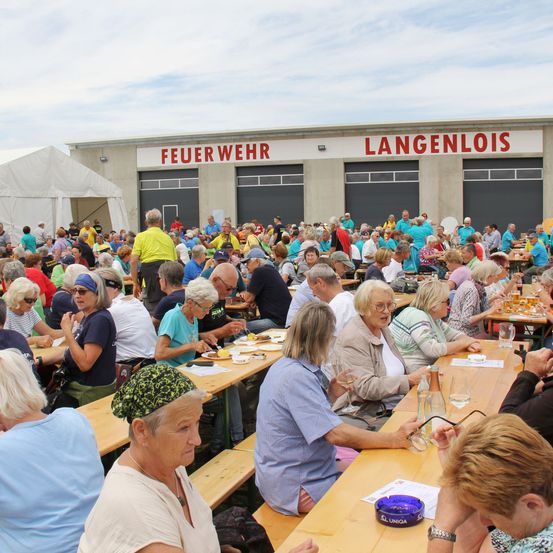 Eine große Gruppe von Menschen versammelt sich vor einem Gebäude mit der Aufschrift 'Feuerwehr Langenlois'. Sie sitzen an Holztischen, essen und trinken. Einige tragen Brillen, Hüte und Sonnenbrillen. Das Gebäude hat ein Zelt links und drei Garagentore.