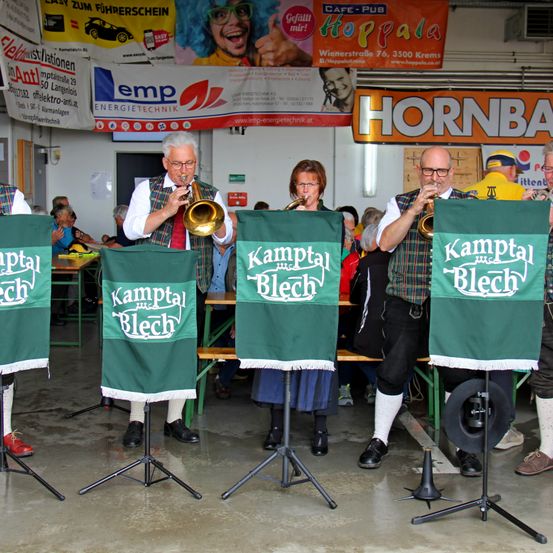 Eine Gruppe von Musikern spielt Trompeten bei einer Veranstaltung. Grüne Banner mit dem Text 'Kamptal Blech' stehen vor ihnen. Hinter ihnen ist eine Menge zu sehen. Es hängen Banner an der Wand.