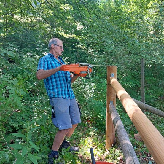 Ein älterer Mann in einem blau karierten Hemd und Shorts benutzt eine orange Kettensäge, um einen Holzzaunpfosten in einem dichten Wald zu schneiden.