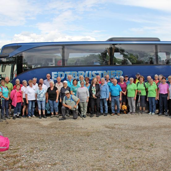 Eine Gruppe von Menschen, einige in grünen Shirts, steht vor einem blauen Bus mit dem Namen 'EDBRUISNER'. Der Bus ist auf einer Kiesfläche unter einem teilweise bewölkten Himmel geparkt.