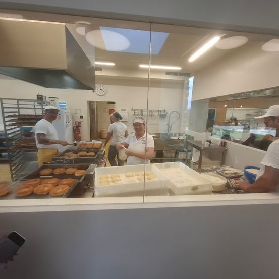 In einer Bäckerei sind vier Arbeiter mit Teig und Donuts beschäftigt. Tabletts mit Donuts sind links angeordnet und Behälter mit Teig rechts. Die Bäckerei hat ein modernes Design mit Glaswänden.