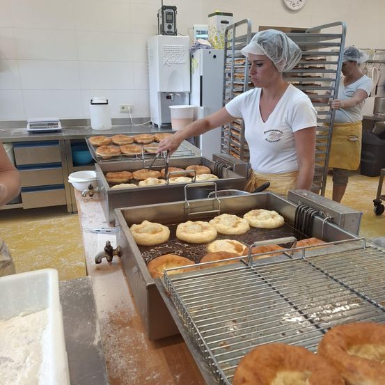 Zwei Frauen frittieren Teig in einer gewerblichen Küche. Eine Frau trägt ein Haarnetz und eine Schürze, während die andere in der Nähe steht. Ein großer Ofen und verschiedene Küchengeräte sind sichtbar.