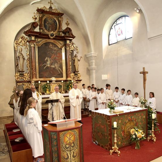 Eine Gruppe von Priestern und Kindern in weißen Gewändern steht vor einem Altar in einer Kirche. Der Altar hat ein Kreuz, Kerzen und Blumen. Der Priester spricht in ein Mikrofon.