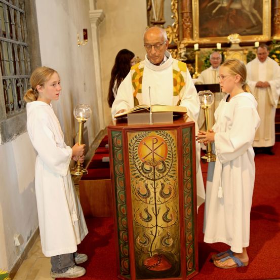 Ein Priester liest von einem Buch am Altar. Zwei Mädchen stehen in der Nähe und halten Kerzen. Hinter ihnen sind andere Menschen in Weiß gekleidet.