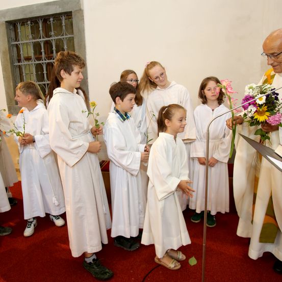 Ein Priester mit Brille steht an einem Pult, umgeben von Kindern in weißen Gewändern, die Blumen halten, drinnen.