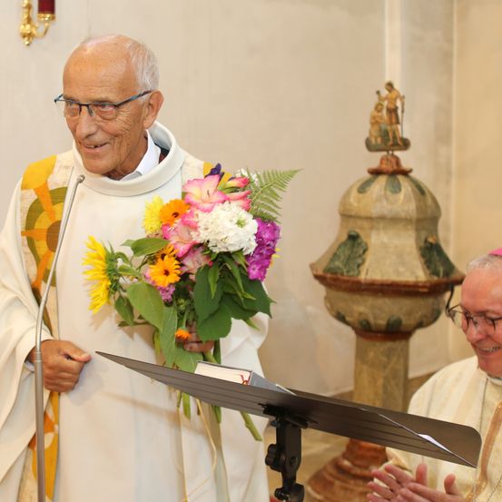 Ein älterer Priester lächelt, während er einen Strauß bunter Blumen hält, und steht hinter einem Mikrofon. Ein weiterer Priester lächelt neben ihm, und sie befinden sich in einer Kirche mit einem dekorativen Steinpfeiler.