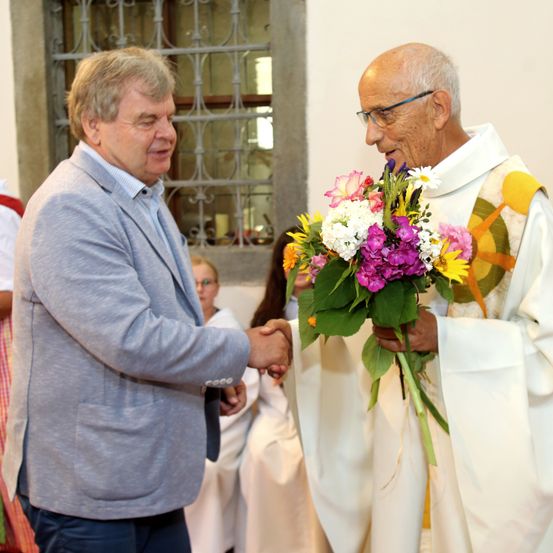 Ein Priester in einem farbenfrohen Gewand hält einen Blumenstrauß und schüttelt einem lächelnden Mann in einem blauen Anzug die Hand. Sie befinden sich in einer Kirche, mit einem Fenster und Personen im Hintergrund.